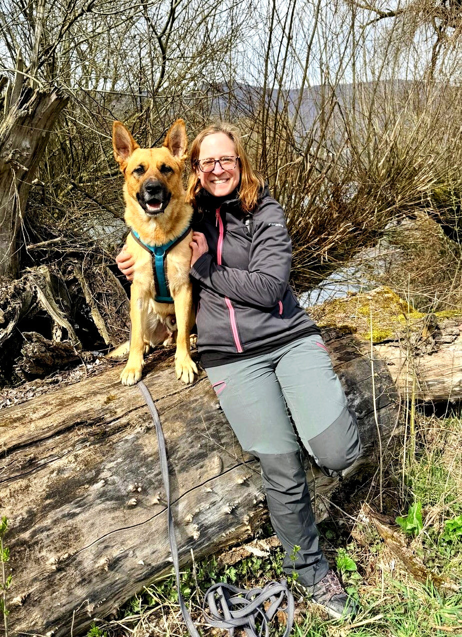 Sabine Müller sitzt mit ihrem Schäferhund Eiven auf einem umgestürzten Baumstamm am Flussufer